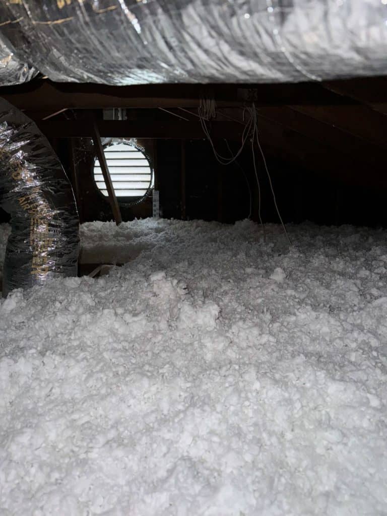 Attic space filled with loose-fill blown-in insulation covering the floor, with visible air ducts and a gable vent in the background.
